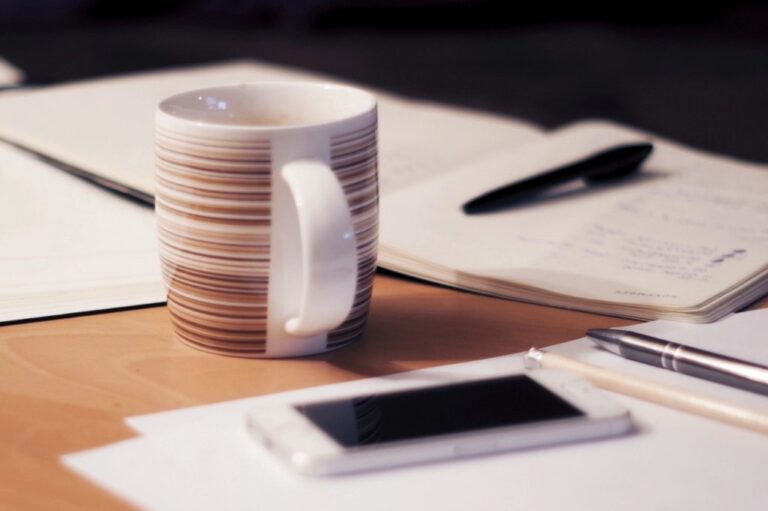 A coffee mug, open notebook and pen on a wooden desk in soft light