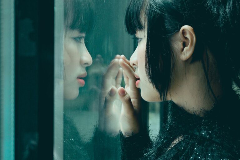 A woman gazes at her own reflection in a window, with two faces visible side by side; identity, self-awareness, dissociation