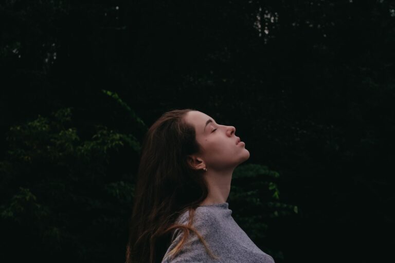 A woman stands in darkness with her face lifted and eyes closed; calm, breathing, nervous system regulation
