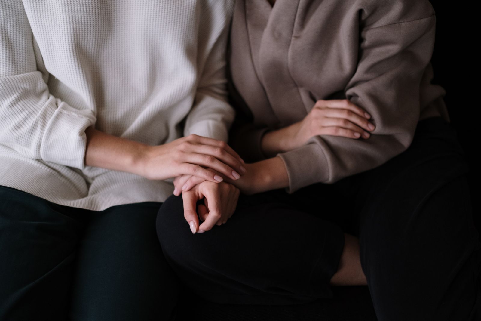 Two people sit side by side, one resting a hand on the other’s hands; comfort, support, being heard