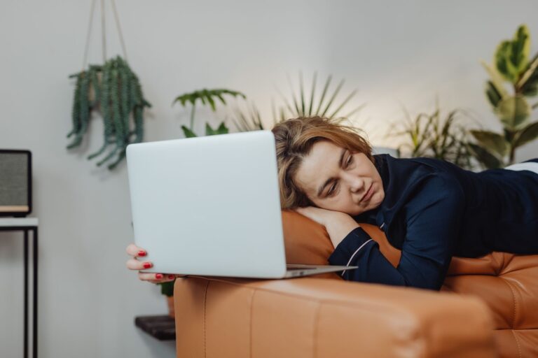 A woman asleep on a couch with an open laptop beside her and houseplants in the background